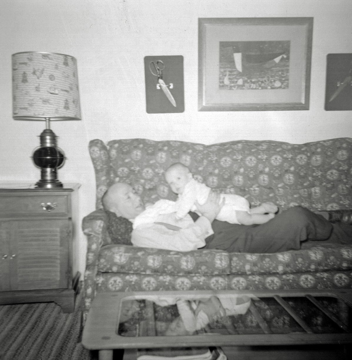 As the sixties replaced the fifties, my mother gave away most of the original mid-century modern furniture and turned to colonial revival to decorate our Levittown house. Here my father Howard plays with my less-than-year-old brother Iden on our new red couch, next to a very sixties record cabinet, faux ship’s lantern light fixture, and in front of a left-over glass and blonde wood  mid-century modern coffee table. Above them is a commercial art print of a duck between a set of Issar’s scissors. My mother treasured those scissors. They were her father, Issar’s, primary professional tool. They are mounted on two green velvet-covered wooden cutting boards, using a brass drawer handle (and gravity) to hold them in place. The way my mother mounted them allowed them to still be used as scissors, though their huge size and weight made them impractical.