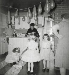 This must have been a big deal party because the woman in the foreground with her hand at her hip is my grandmother, Marie. She lived two states away from Levittown, Pennsylvania, in New York City. She did not visit often.

Leaning against the sink, with her arm also on her hip, is Dorothy Neil, our next door neighbor. My mother, Arlene, is the center adult.  What she is doing at the counter, I don’t know. But this picture makes it clear that it is her arm getting something from under the sink in last week’s picture.

I am guessing that the crouching child is me because she seems to be setting the not-table with plastic candy baskets. I can’t imagine a guest doing that. This shot also makes me question the date. Standing up, these girls look bigger than nursery school. I would guess they were kindergartners. But they can’t be because the Neils did not live next door to us in 1960. They did in 1959 and 1961.  So these must be first graders, making it my seventh birthday.

