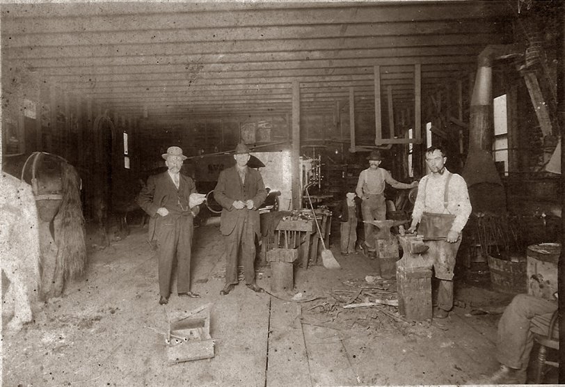 Inside shot of a blacksmith shop. Don't know place it was taken. Appears to have a car from the twenties in the back of the shop. Photographer unknown.