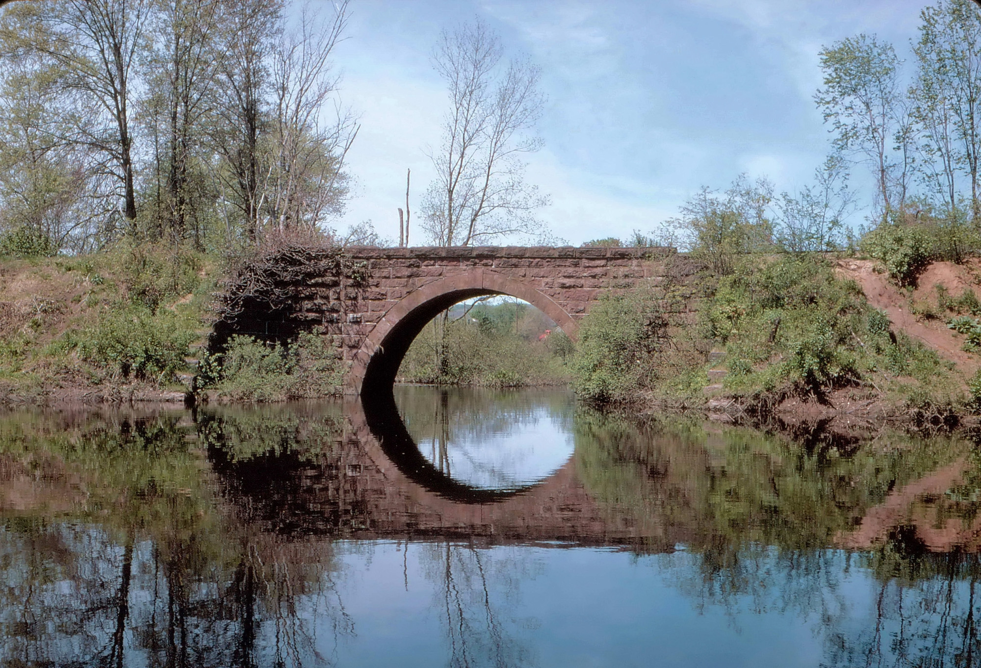 The "Canal Line" railroad, so-called, was constructed in 1847-8 from New Haven, Connecticut to Northampton, Massachusetts to replace the failed Farmington (later New Haven - Northampton) Canal, running over roughly the same course as the canal. The brownstone arch spans the Tenmile River in Cheshire, Connecticut.  In 1987 Interstate 691 was built though this site and the bridge was replaced with a modern banality. Numerous railroads have owned the old line over the years and all rail traffic on this part of the line ceased very soon after its replacement. A rail-trail extension through this area is expected. I took this picture on June 4, 1967 when I was 16. Kodachrome, of course! View full size.