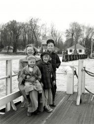 One of our annual trips from New Jersey to Florida circa 1951-52. This was pre-Interstate days and we traveled U.S. 17, 301 or sometimes A1A. Not sure which ferry this is. With Mom, sister and older brother. View full size.
Nanticoke River at Seaford, Delaware?The Woodland Ferry lies on a possible route between New Jersey and Florida via Delaware, Maryland, and Virginia. It is a cable ferry. Here is a link to photo of the 1938 version that was replaced about 1961. The chain does not show in the Shorpy photo but might be explained by photographer standing with back to cable and operator shack. A second ferry ride awaits our travelers when they reach the Chesapeake Bay. 
My family took the Chesapeake ferry ride in 1947 when we traveled from Jacksonville, Florida to visit my aunt in Trenton, New Jersey. I remember the Chesapeake crossing because my four year old brother and I held our three year old brother over the railing so he could see the water. 
http://capegazette.villagesoup.com/media/Common/0/5B/23510/t600-pg_7_pic...
Looks Like The Fort Gates FerryAnd it's still in service today. This is almost certainly the Ft. Gates Ferry which has been operating since the 1850s, crossing the St. Johns River south of Palatka, Florida, north of Lake George, between the Fort Gates Fish Camp and Salt Springs in the Ocala National Forest. It is the oldest ferry still operating in Florida.
This ferry boat consists of a 1914 steel barge, propelled by a converted 1910 Sharpie sailboat that is permanently connected to the barge.
The last time we took it the toll was $10, well worth it.
[It certainly seems like a possibility, but how do we know it's the Fort Gates ferry? - Dave]  
Not FloridaThe Fort Gates ferry is reached by 12 miles of dirt road on West and 1 mile of dirt road on East making this an unlikely route for a vacationing family. The ferry in the photo has a deck mounted air vent suggesting an on board engine. The built up area on the shore is also questionable.
The ferry Jean LaFitte at the Mayport crossing of the St. Johns River in the 1950s was a much larger vessel with enclosed deck. The vessel in the photo is no match for currents running at mouth of the St Johns River.
I grew up in Jacksonville, and as much as it pains me, I vote against Fort Gates.
Chilly WeatherJudging from the winter hats and coats being worn, my guess might be that the location is somewhere to the north of Florida. 
(ShorpyBlog, Member Gallery)