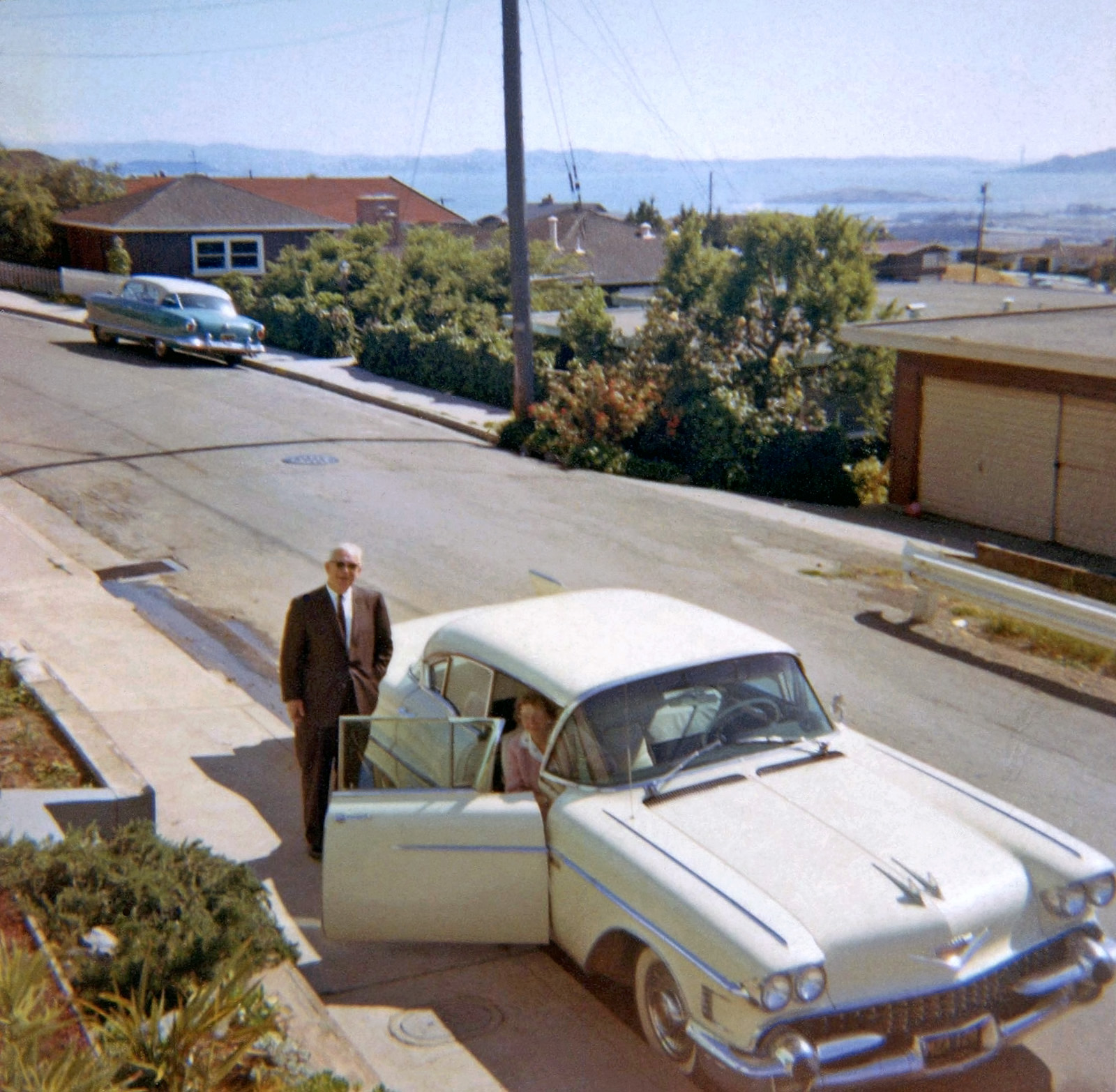 My mother's parents are seen here getting ready to drive home to Sacramento after a weekend visit in 1964. We lived in the San Francisco East Bay hills for over 20 years and this house had a wonderful panoramic view of the Bay.  San Francisco can be seen in the left distance as can the Golden Gate Bridge in the distance at right.  Grandpa's car was a 1958 Cadillac - he was a Cadillac man for years. The car down the street is a c.1954 Nash Ambassador.  As a toddler I would stand out on our deck and stare out at that incredible view. View full size.