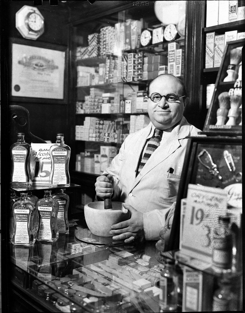 Here is my paternal grandfather, William C. Dembling, behind the counter of his drugstore at 60 W 8th St in Manhattan NYC, around 1935. I have the feeling this picture may have been taken by my father, but I have no way of knowing for sure.
A full-fledged pharmacist and medicinal chemist, "Doc" Dembling was a well known and well loved fixture of the Greenwich Village of the 1930s. He treated for free many a hangover, and a few more serious ailments, for lots of the penniless artists and other characters (many of them nefarious) who roamed the Village during the depths of the Depression. His shop also included a busy soda fountain and sandwich counter.  
Doc was also known to have done a little...er...private manufacture of "spirits" during Prohibition. He was quite a character. My father resembled him strongly but kept all his hair until his dying day. 
I have always regretted never meeting my grandfather, but alas he passed away in 1941, quite a few years before my birth. He was by all accounts a gentle, kind and generous soul. I intend to post a few more pics of his extremely well stocked drugstore; a classic small New York business of the 1930s. View full size.
