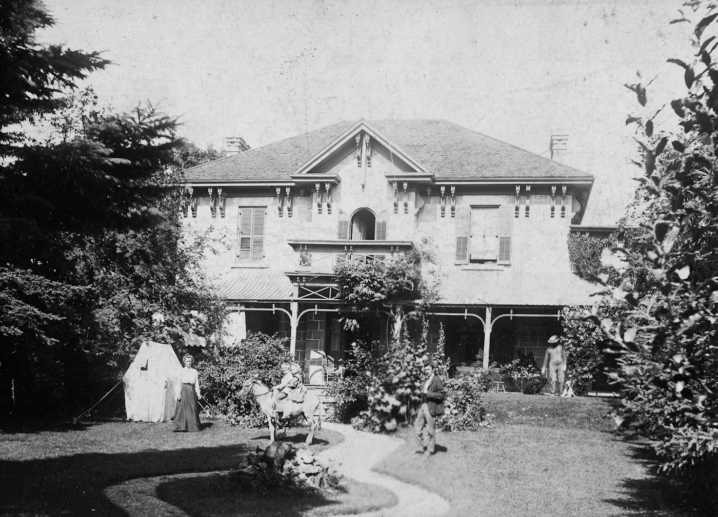 This is the house of Dr. George Devey Farmer, in Ancaster, Ontario, taken about 1905. In the photo are Dr Farmer, his wife (and first cousin) Eleanor Shelton Farmer, and two of their four children - George Richard Devey Farmer and Margaret Alice Devey Farmer - sitting on Balaam the donkey. To the right is Collinson, with the dogs.  

The house was built about 1873 for Dr. Richardson, and still stands at 343 Wilson Street, Ancaster, although shorn of its veranda and fountain.  

The Farmer family (Dr. Farmer's grandfather) arrived in Ancaster in the 1850s, having arrived in Canada twenty years before, from Brockton House, near Shifnal, Shropshire. All their possessions were loaded into a chartered 430 ton sailing ship - the Kingston, out of Liverpool.  They included 42 packing cases of furniture, all of their animals, and many of their tenants.  There were 45 people in addition to the family.  The voyage took 51 days.

Dr Farmer served in World War I in the Wentworth Medical Corps, and served the village and rural area for many years as doctor.  He owned the first automobile in Ancaster, a Pope, from 1902. View full size.