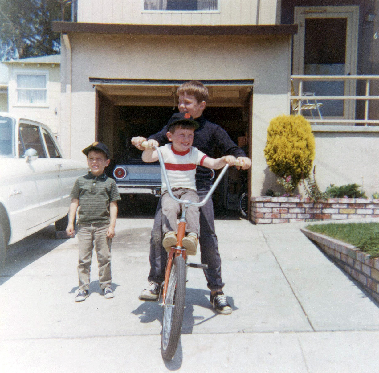 There I am in the white shirt and Giants cap riding with my friend Robert.  He taught me to ride a bike after getting tired of hauling me around like this.  One day he got off the bike and gave me a push and I was solo for the first time!  Next-door neighbor Ray stands by as we prepare to launch.  Dad's 1962 Ford Falcon at left was soon traded for a new '66 MG Midget and the Midget was soon traded for a new '67 Austin Healey 3000.  Mom's new '66 Ford Falcon resides in the garage.  This was our house on Claremont Avenue in East Richmond Heights in the San Francisco Bay Area. View full size.