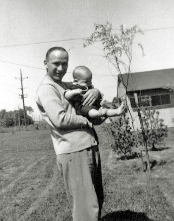 I am the six month old bundle of squirms in my father’s arms as he stands behind his first house in Levittown, Pennsylvania during September 1954.	Photo was taken on a (then newly purchased) Argus 75 box camera by my mother. Scanned from a print in tribute to my father, Howard, who lived from September 1924 through June 2014.