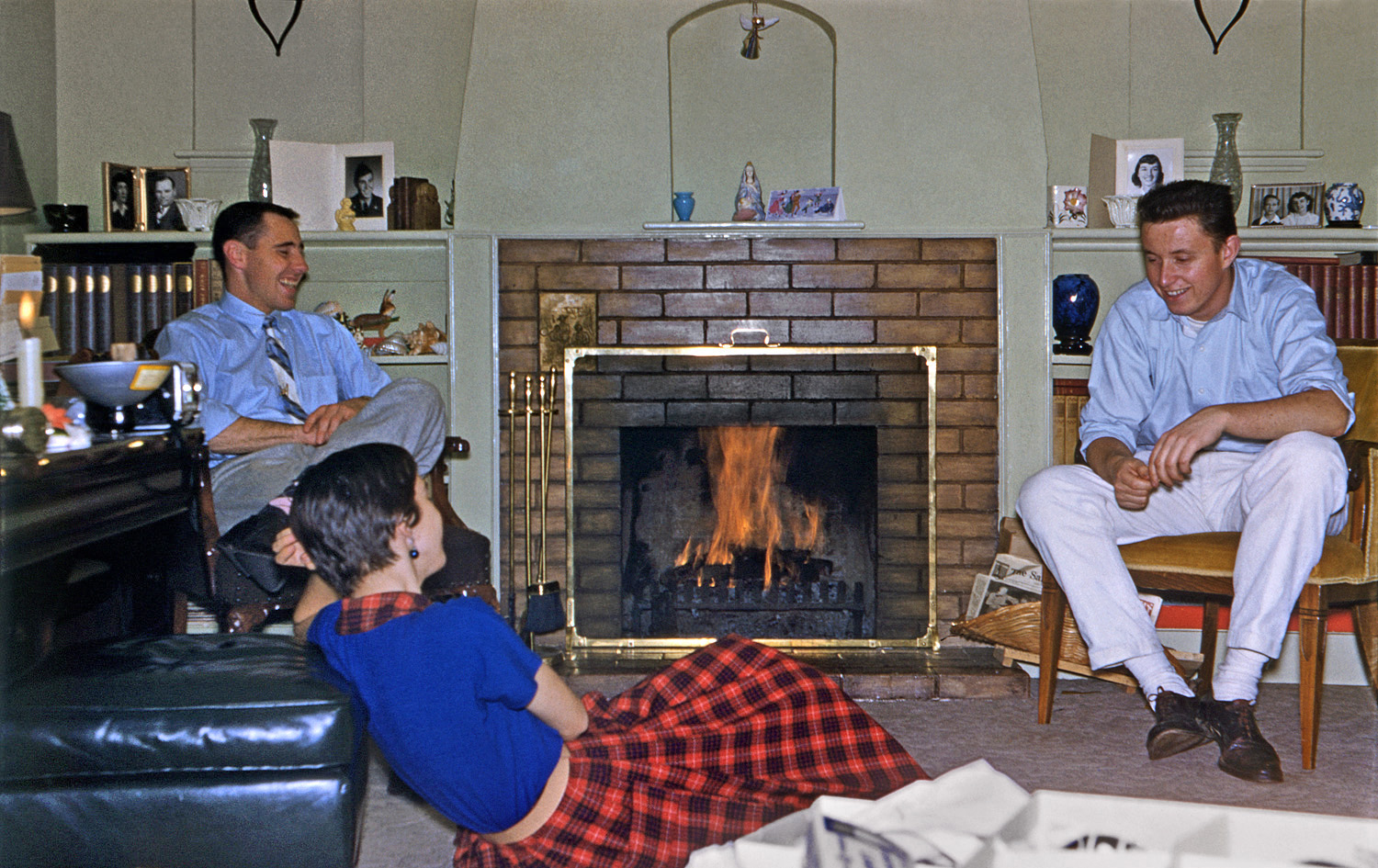 December 17, 1955. Tony W.'s baby shower photo, with the fireplace and shelves full of photos, books and bric-a-brac, was so exceedingly redolent of 1950s living rooms in general, and ours in particular, that I couldn't stand it, so I dug this one out, showing our fireplace and shelves full of our photos, books and bric-a-brac. That's my sister, with her future husband on the right, and my godmother's son on the left. That's him in Army uniform in the photo on the shelf above. My brother, bless him, wrote the actual date on the slide mount, but we can tell it's near Christmas by my sister's home-made angel ornament hanging in the niche as well as the box of tree ornaments in the lower right foreground. Another point of interest is the candle burning at the extreme left. It's functional, not ornamental; this was taken during a power outage occasioned by the record Northern California December 1955 storms. Also on the table next to the candle is my sister's Kodak Duaflex camera with flash attachment. View full size.
