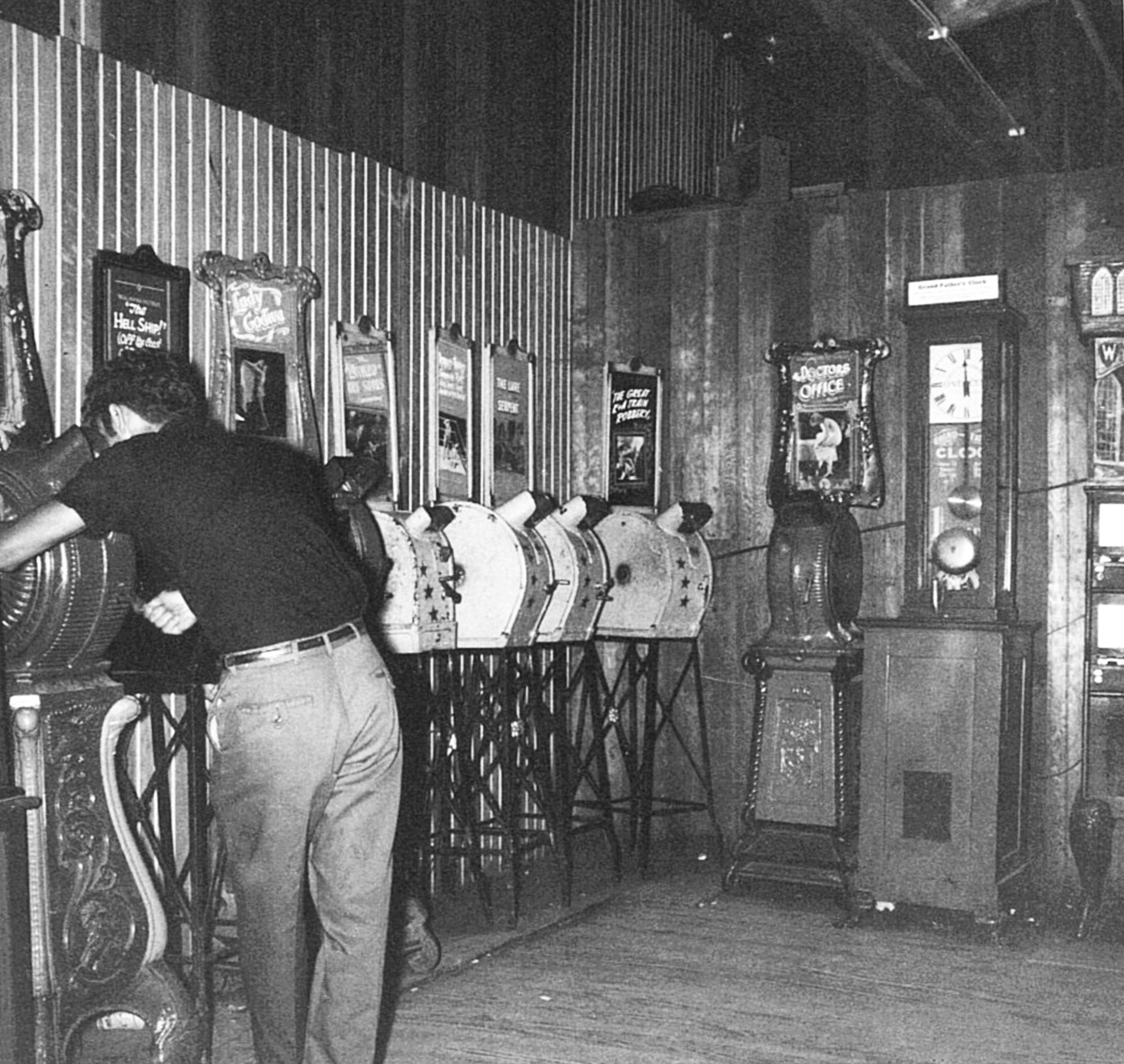 From an arcade in Upper Dauphin County, PA. This picture dates to around the mid to late 1950's even though the machines are much older. Just shows that they were built to last. 

The first 8 machines, beginning on the left and going clockwise, are a variety of Mutoscope movie viewers. A couple cast iron "Clamshell" models, (c.1895), a rare "Indian" model (c.1895) that the man is using, and a few all tin models. In the right half of the picture is a c. 1925 Gatter Novelty Company "Grandfather Clock Strength Machine". To the far right, and cut off is an Exhibit Supply Card vendor from 1930. These machines are highly sought after by collectors. View full size.