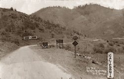 This is a postcard dated 1928 showing the Pacific Highway on the Calif.-Oregon border. Most likely Interstate 5 now. Looks like it's slow going on that gravel. No 65 mph on this road.  
Photo LocationIf the photo was taken on the Pacific Highway would that not now be Highway 101 on the coast? I-5 is some distance inland. That would place the location about 4 0r 5 miles south of Brookings, Oregon
pacific highwayIt's not the pacific coast highway, anyway I don't think all the signs are readadle at the resolution that the photo is posted at but one of them says Siskiyou county. 
Pacific HighwayWikipedia article on the Pacific Highway.
Speaking of resolution, Kev, when you save your scans as jpegs you should choose "large file size" or "high image quality." Often this is shown as numerical scale from 1 (low quality) to 12 (high quality). All of your jpegs have a quality level of 3, which is why they are kind of fuzzy and pixelated.
Re: Pacific HighwayNobody said this was the Pacific Coast Highway. It's the Pacific Highway, a completely different route.
Oops.Pardon my conclusion jumping.  Pacific Highway, it is, and that would make it Highway 99.  
Don Hall
Yreka, CA
Please post a higher resolution image.It's definitely not the Pacific Coast highway if it says Siskiyou County.  Looks like it might be the border of old Highway 99, although the vegetation doesn't look quite right.  Would love to see a higher resolution picture so I can check out the details.
Don Hall
Yreka, CA
Pacific HighwayThe old stage coach road is still visible as it crosses old Hwy 99 and I-5. This picture is probably that same road. I'll bet that sign next to the car tells you a list of things you aren't allowed to do or possess in California.
US 99Looks to me like Jefferson Road, Old UD 99, that runs to the east of I-5 / SR 99.
LocationThis may be the location where this photo was taken. It's on the Pacific Highway/Route 5 on the border of Oregon and California. The hills in the background look the same in both photos.  The  California/Oregon stateline sign is in approximately the same place as in the old photo. You can't see it in this new photo but it's on the right side, just out of the frame.
Sign and DateThe sign to the right of the auto reads: "Visiting Motorists / the California State Automobile Assn. / Is At Your Service / Non-Resident Registration Certificates / Required by State Law. Tourist Map, General / Road Information and Digest of the State / Traffic Laws Supplied Without Charge."
I can only read the top line of the sign the auto is partly obscuring: "Non-Resident Motorists."
The earliest postmark I've found on this postcard is July 1926.
When the photo was taken the road was indeed gravel on the California side, but the Jackson County, Oregon segment had been paved for ten years. You can see the end of the pavement in the foreground.
Another crossingThis is near the town of Hornbrook California. My father was traveling through this area in 1923 and his Model T burned out a clutch. He had the clutches replaced in Hornbrook. I am attaching a photo of where he crossed the state line and am not sure where it was. I know it must have been near here, or possibly down river near Happy Camp. He said he had to back up some hills in reverse to get to Hornbrook. The boarded-up garage was still there in 1970 when we drove through there.
(ShorpyBlog, Member Gallery)
