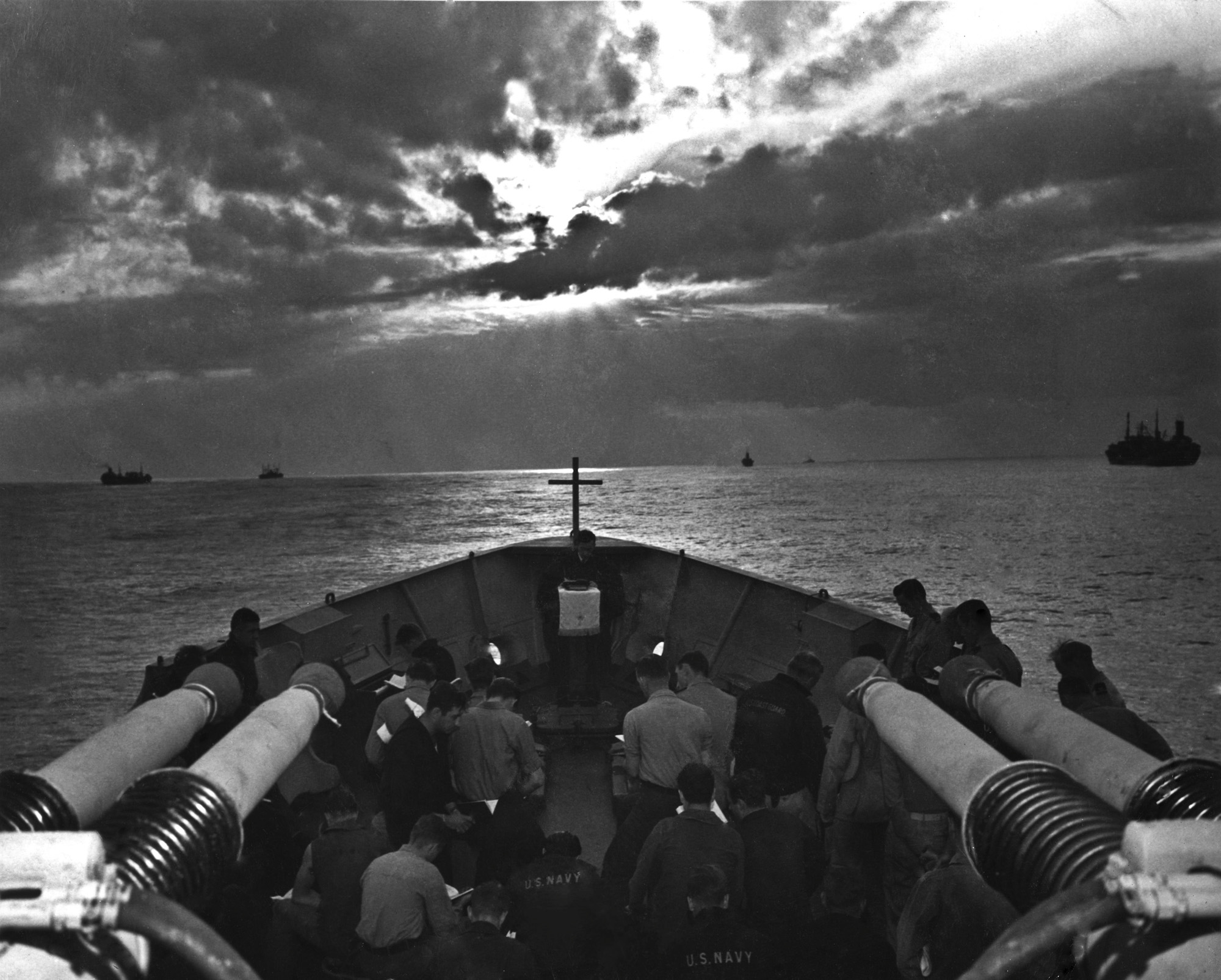 This photograph was taken Easter Sunday, April 9th, 1944 aboard the USS Duane. This photograph was scanned from my grandfather John "Jack" Baker's Warbook. If you look closely, you will see a cross at the bow of the boat, behind a chaplain reading from a bible. I find this photograph particularly beautiful, with the sun rising between the clouds across the front of the ship. There is a stark contrast between the peacefulness of the men reading, and the reality of the massive guns pointed above their heads. This photograph wasn't marked, but it was likely taken by Dale Rooks. There are no identifiable men in the photograph. View full size.