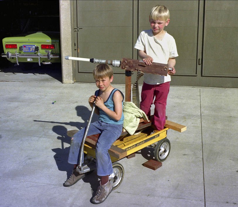 This is my brother Paul (right) with a friend, Danny Bergman, who lived four or five houses down the block.  It was taken in 1973 on the driveway in front of our house in Huntington Beach, California.  I built the "machine gun" for them from spare pieces of PVC, wood and assorted hardware that were lying around in the garage.  Next, I nailed a belt of machine gun ammunition to the block of wood and voila, a "machine gun" ...  I used to go hiking with my father up in the dusty hills of Camp Pendleton where I collected spent rounds and belt links from the range.  (At the time, I had no clue that I would end up hiking those hills again in 1979 and 1980 as a Marine myself!  Or did I?)  When I got home, I would reassemble the brass casings with the belt links and make lengthy ammunition belts.  In the late sixties and early seventies I always had an ammo can with ten or so feet of belted machine gun rounds. My friends and I played "Army" a lot back in the sixties.  Anyway, my brother and his friend would go around playing "Rat Patrol" and blow away all sorts of imaginary suburban enemies.  In the garage is my parents 1972 Fiat 124. I never got to drive that thing but it was a dog of a car from what I remember. They ended up replacing the Fiat with a light blue 1975 Volkswagen Rabbit.  I learned to drive in the 4-speed manual Rabbit while I had my learning permit.  I got my license on the day of my sixteenth birthday and drove the Rabbit until 1976 when it was sold after being involved in an accident.  I was not driving it at the time! Scanned from the original 110 negative. View full size.