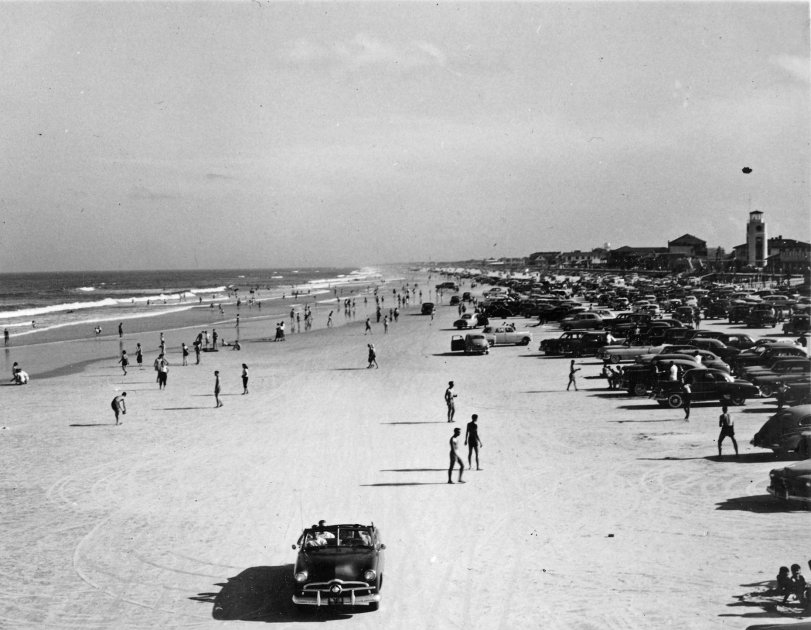 Cars drive on the beach in New York, San Diego or Virginia. The tower on the right does not appear in any other commonly available photos of New York-area beaches, so I could be completely off base.

It's from the early 50s, as best I can tell from the makes of the cars depicted, and the pile of old family photos in which I found it. My dad was stationed at NAS Miramar (now MCAS Miramar) and Naval Station Norfolk during the Korean War, which is why there's an outside chance it's a beach in the San Diego or Norfolk area.
