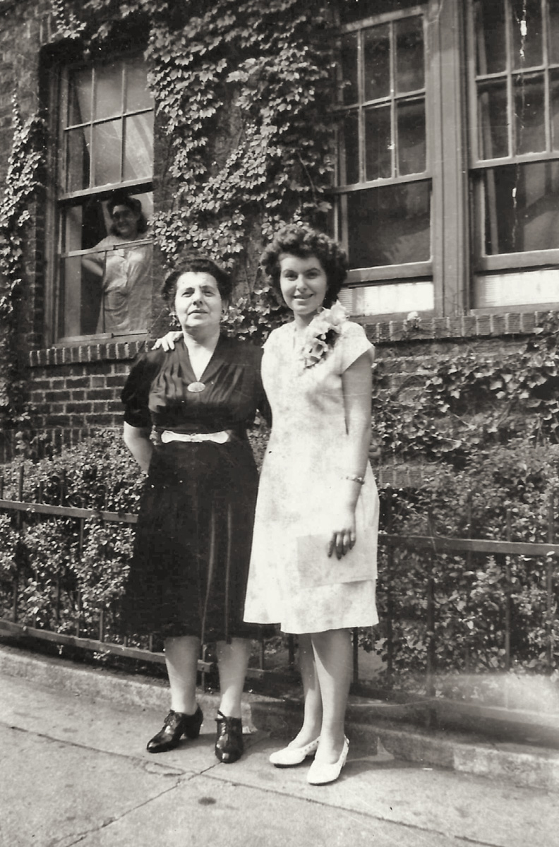 My then 14-year-old future mother poses with her mother in front of their Brooklyn, New York apartment to commemorate my mother’s junior high school graduation, while a neighbor watches what is going on from her window.