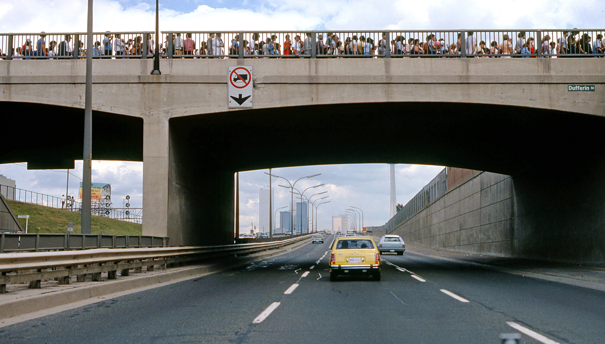 This is the second of three submissions of a 1979 Labour Day trip to Toronto on the QEW, documented from the driver's seat. Here I'm approaching an overpass with a line-up of people. They're waiting to enter the CNE (Canadian National Exhibition) by way of the Dufferin St. Gate. The CNE has been held for well over a hundred years on the waterfront of Toronto, and attracts hundreds of thousands annually at summer's end for two weeks. Downtown Toronto and the CN Tower can be seen in the distance. View full size.