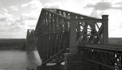 Unknown location, but a pretty big bridge. Any guesses where this is/was? From my negatives collection. View full size.
The Quebec RR bridgeI believe that is the Quebec RR bridge, completed in 1919 after the first attempt/design failed and collapsed into the river.
Pont de QuebecCorrect ID, rnold.  And taken from the same angle as the shot below.  Re mhallack's "pretty big bridge": it is the longest cantilever bridge span in the world.  It actually failed twice during construction: the first time in 1907 (75 workers killed), then again in 1916 (13 killed).
Much like the Forth Bridge in EdinburghWhen I first saw the picture I thought it was the Forth Rail Bridge in Edinburgh, Scotland, although the structural members looked a bit different. The main upright members of the Forth Bridge are tubes, rather than square or rectangular. 
It turns out that the similarity isn't coincidental - one of the engineers appointed to do the 1916 re-design after the original design failed in 1907 was Maurice FitzMaurice from Britain, who had worked on the Forth Bridge.  
(ShorpyBlog, Member Gallery)