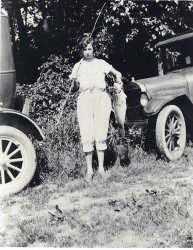 This is my wife's grandmother holding up her catch (looks like a big carp!) after a day of fishing with a cane pole in 1917 on the Little Miami River near Cincinnati, Ohio.  She was 17 years old at the time and lived until 1969. The cars look like a couple of Model T Fords. Those old "flivvers" would go anywhere! View full size.
1917You know, the car to the right is a late "T", a 1926 or 1927, which had different design features than all those that came before it. Most notably, the gas tank was moved to the cowl, and there is a flap or door in front of the windshield on these to add gas. They also have a headlight bar, which the pre 26's did not. The rear car gives away the fact the photo cannot be 1917. Add to that the observation that the paint is no longer shiny, and the fender is banged up, and I'll bet this photo is of a car at least 3 or 4 years old. So the photo must be the 1930's! 
(ShorpyBlog, Member Gallery)