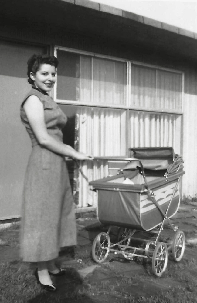 One month after my birth, my mother Arlene poses for a series of snapshots taken by my father in their back yard with the new baby, or in this case with the new carriage they bought to use with me.
