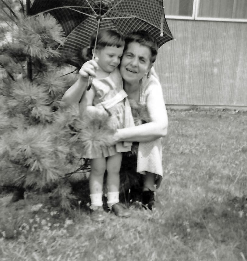 Grandma Marie came to visit and brought three year old me a red plaid umbrella as a present. Then my mother wanted to take some pictures of me with her mother. So we went into the back yard. Of course I took the brand new umbrella with us. No matter how much Marie pleaded with me to set the umbrella aside, just a minute, for the picture, I would not. She even tried to bargain a compromise: some pictures with the umbrella, some without.
No deal.
I was hanging on to my new present, no matter what. Photo was taken by my mother. Scan was made from a print.
