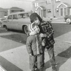 I am the three-year-old child overly bundled up in the red snow suit and trying to get away from the older boy (who was a son of my father’s sister). He kept pushing my cheeks together to give me a funny face, and I kept trying to stop him. But the star of this photo is a 1956 Chrysler parked on his New York City Street that we happen to be blocking. Why, oh, why couldn’t we have been standing three feet further back to give us a better view of the car?
