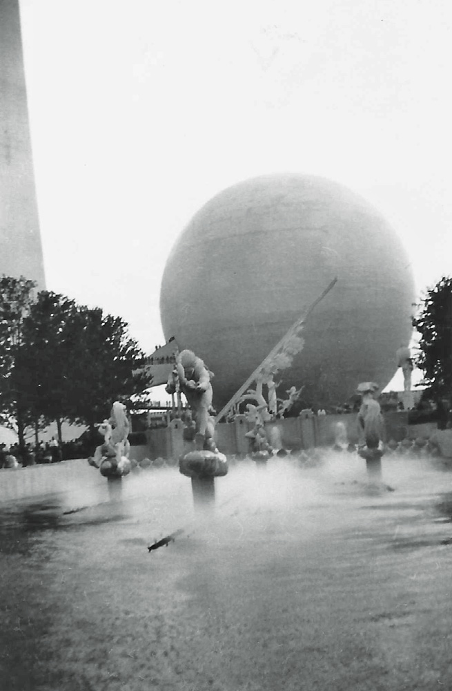 Statues in the misty Lagoon of Nations stand in front of the Trylon and Perisphere at the 1939 New York World's Fair, dwarfing the tiny people waiting in line to enter the exhibit inside the ball-shaped Perisphere.