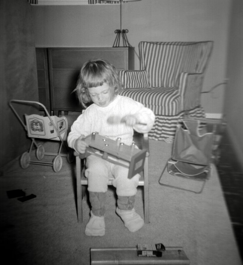 One of the things you find out in editing your family pictures for sharing on Shorpy are the strange things you never thought of when you were growing up. My family seems to have had “a thing” about carriages and strollers, both real and for play. In this picture, which shows me in the foreground using my potty chair as a regular chair and playing with a pound-a-peg bench, there are two play strollers behind me. A tin lithographed one is standing upright, and a steel frame and fabric one is turned over on the floor.  Other pictures show me climbing into a carriage, and wheeling it on the sidewalk, and wheeling a dog in the stroller that is on the floor in this picture.
I remember a fourth woody wagon carriage from the 1940's that I have not found a picture of. And then there are the numerous pictures of me or my brother in carriages or strollers, and even one of my family using one with my brother at the kitchen table in place of a high chair.  Why did I have so many strollers and carriages and why are there so many pictures including them? I have no clue.
