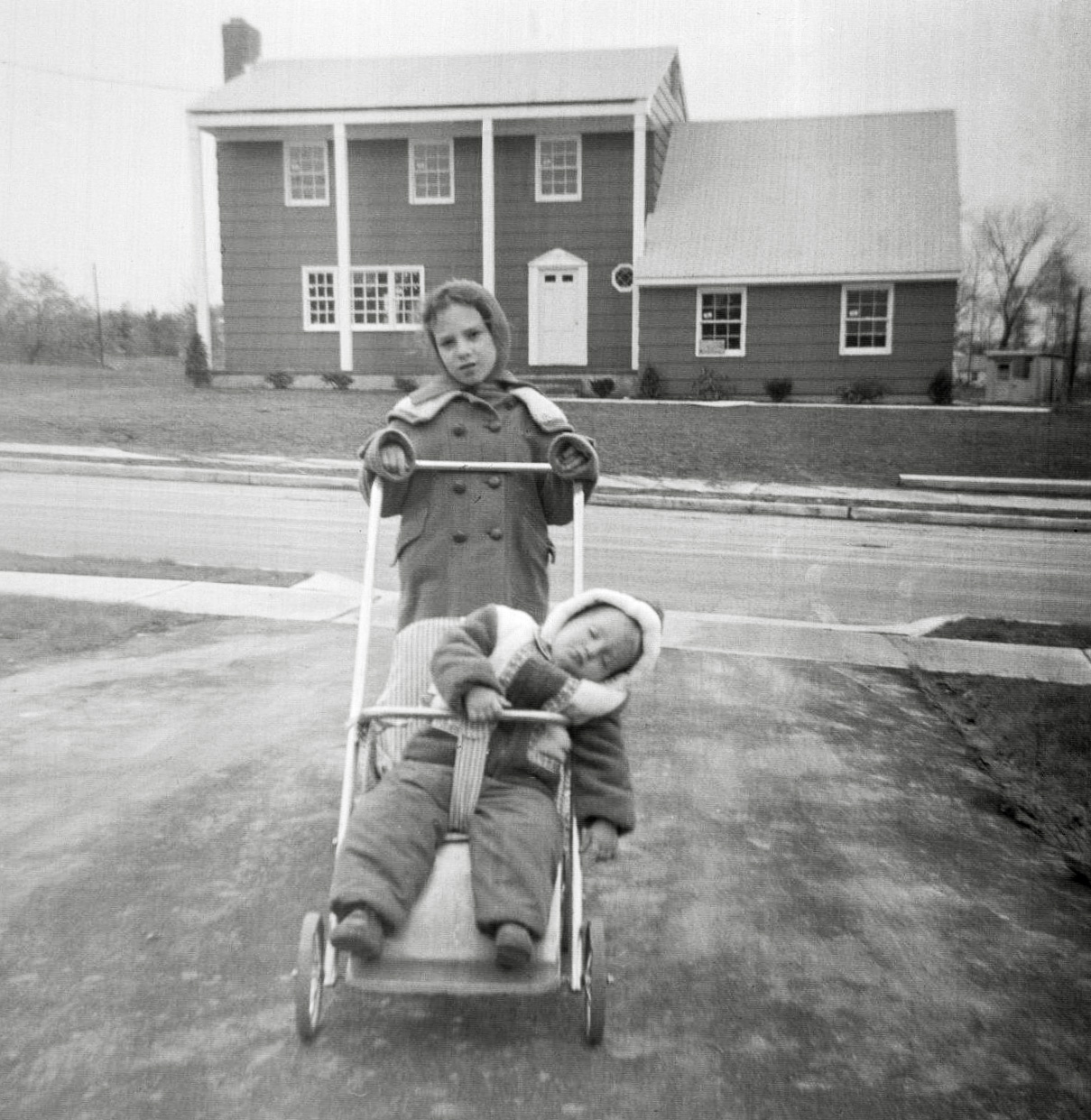My mother thought it was funny how my baby brother fell asleep in his stroller, so she posed me with it and took this picture. The date is early 1963, around six months after we moved into our new Princeton, New Jersey house. 
Like in Levittown, my family were the original owners, though unlike Levittown, these were custom-built homes. You selected your lot, then selected a blueprint from the library of blueprints that the builder had. The blueprint was just a starting point. You then edited those basic plans. In our case we added footage to the garage to fit our 1960 Edsel, because the garage, as drawn in the plans, was shorter than the car. Closet configurations were another thing my mother totally customized.

The just-completed, though not yet occupied house across the street stands starkly in the background with its not-yet-grown landscape and bare lawn area. As for me, I seem to be stoically putting up with my mother’s photography.”Say cheese” was not a phrase in my family’s vocabulary.
