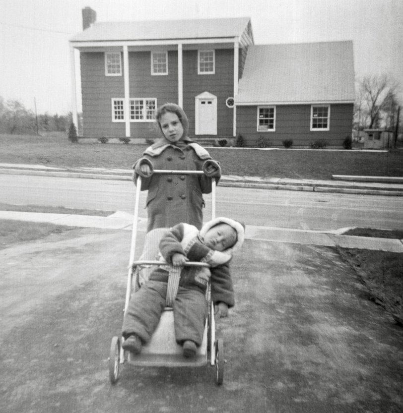 My mother thought it was funny how my baby brother fell asleep in his stroller, so she posed me with it and took this picture. The date is early 1963, around six months after we moved into our new Princeton, New Jersey house.
Like in Levittown, my family were the original owners, though unlike Levittown, these were custom-built homes. You selected your lot, then selected a blueprint from the library of blueprints that the builder had. The blueprint was just a starting point. You then edited those basic plans. In our case we added footage to the garage to fit our 1960 Edsel, because the garage, as drawn in the plans, was shorter than the car. Closet configurations were another thing my mother totally customized.
The just-completed, though not yet occupied house across the street stands starkly in the background with its not-yet-grown landscape and bare lawn area. As for me, I seem to be stoically putting up with my mother’s photography.”Say cheese” was not a phrase in my family’s vocabulary.
