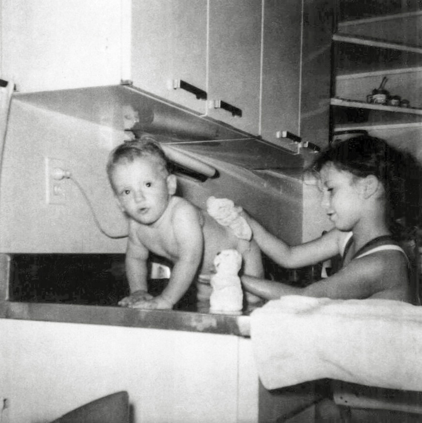 This June 1962 photograph was taken by my mother and shows me and my brother Iden in the kitchen of our home at 110 Dogwood Drive, Levittown Pennsylvania. It is an entirely posed picture of eight-year-old me replicating a 1955 photo where my father washed me in the same sink. My brother really was washed in the kitchen sink when he was a baby, just like I had been. But I did not do the washing. My parents did. I think it was me who wanted there to be matching pictures for me and my brother, and so suggested to my mother, when she was washing him, that she get the camera and we create the picture again. My father was probably at work, which is why I got to be the washer.