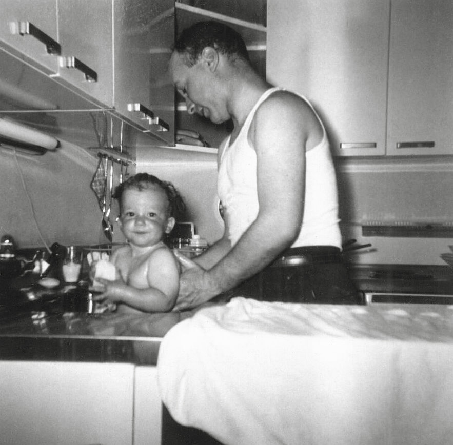My mother took this photograph of my father washing me, in the kitchen sink, some time in 1955. That kitchen was one of many hundreds exactly like it in the Dogwood section of Levittown Pennsylvania. Both the sink and counter were stainless steel and the cabinets where white painted steel with stainless handles. Though it is hard to see the electric oven in the background, every kitchen got the same GE electric oven/range combo and refrigerator when they purchased the house. My parents added the work counter (on which my father has placed a towel, for drying me after my bath). Otherwise this kitchen is unchanged from when the house was built in 1953. 
