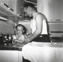 My mother took this photograph of my father washing me, in the kitchen sink, some time in 1955. That kitchen was one of many hundreds exactly like it in the Dogwood section of Levittown Pennsylvania. Both the sink and counter were stainless steel and the cabinets where white painted steel with stainless handles. Though it is hard to see the electric oven in the background, every kitchen got the same GE electric oven/range combo and refrigerator when they purchased the house. My parents added the work counter (on which my father has placed a towel, for drying me after my bath). Otherwise this kitchen is unchanged from when the house was built in 1953. 
Little cutieOh my how cute you were. My grandmother updated her 1906 home with modern steel cabinets in the 50's. I liked them except she always knew when we were in the cookie drawer. 
(ShorpyBlog, Member Gallery)